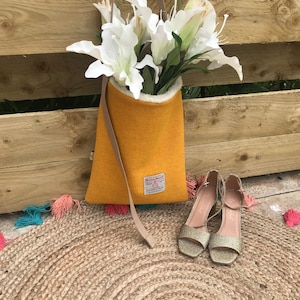 May include: A mustard yellow corduroy tote bag with a white faux fur lining and a brown leather strap. The bag is hanging from a wooden fence and has a white flower bouquet inside. The bag has a small label that says "Holly & Bear".