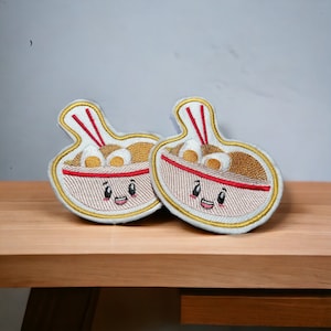 Peut inclure: Deux patchs brodés en forme de bols de ramen avec des visages souriants et des baguettes rouges. Les patchs sont sur une surface en bois brun clair.