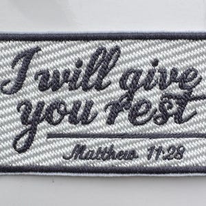 Op de afbeelding: Geborduurde patch met de tekst "I will give you rest" in een cursief lettertype, met "Matthew 11:28" eronder. De patch heeft een grijs-witte achtergrond met een donkergrijze rand.