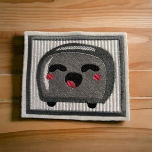 Könnte beinhalten: Ein grauer gestickter Aufnäher mit einem lächelnden Toaster mit roten Wangen. Der Toaster ist schwarz umrandet und auf einem weißen Hintergrund mit grauen Streifen platziert.