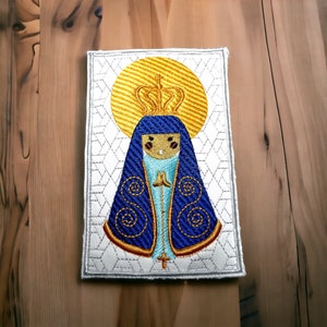 May include: Embroidered patch depicting the Virgin Mary in blue and gold, wearing a crown and halo. The image is set against a white background with a geometric pattern.