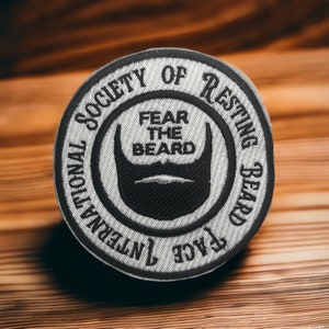 Puede incluir: Un parche bordado blanco y gris con el texto "International Society of Resting Beard Face" y "Fear the Beard" en un círculo alrededor de una silueta de barba negra.