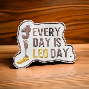 May include: White embroidered patch with a brown outline. The patch features a cartoon leg with a prosthetic limb and the text "EVERY DAY IS LEG DAY."