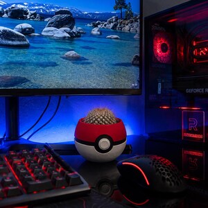 May include: A desktop setup featuring a red and black gaming keyboard, mouse, and a computer monitor displaying a scenic lake view. A red, white, and black Pokeball-shaped planter holds a cactus. The computer case has red internal lighting.