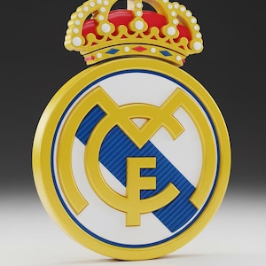 May include: A 3D render of the Real Madrid club crest. The emblem features a gold circle with a blue and white interior, and a gold crown at the top. The letters "M" and "F" are visible.