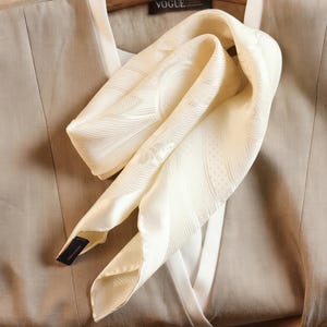 May include: Ivory-colored silk scarf with a subtle patterned design, draped over a beige garment. The scarf has a textured surface and a label with the word "VOGUE" is visible. The scarf is a fashion accessory.