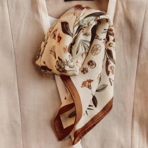 以下が含まれることがあります： 茶色、赤、緑の花柄が特徴のクリーム色のシルクのスカーフ。スカーフは茶色の縁取りで、薄いベージュのトップスにドレープされています。「VOGUE」の文字が木製のハンガーに見えます。 サイズ: 約70cm x 70cm。