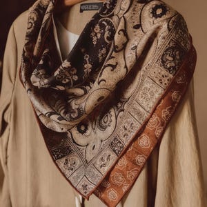 May include: A silk scarf with a paisley pattern in shades of brown, tan, and black. The scarf has a border with a rose design in a darker brown hue. The scarf is draped over a tan blazer hanging on a wooden hanger.
