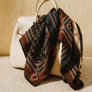 Könnte beinhalten: Eine cremefarbene Lederhandtasche mit einem gemusterten Schal. Der Schal hat ein sich wiederholendes Design aus schwarzen, braunen und goldenen geometrischen Formen und Kettengliedern. Der Schal ist aus einem seidigen Material.