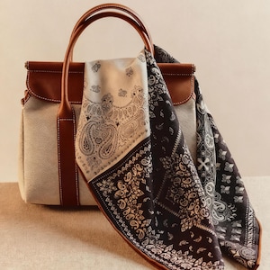 May include: A beige tote bag with brown leather handles and trim. Two paisley patterned bandanas, one black and white, the other cream and black, are draped over the handles. The bag has a structured design.