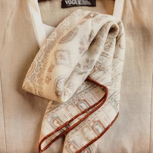 Puede incluir: Un pañuelo crema doblado con un estampado de cachemira y un borde marrón descansa sobre un chaleco beige. El chaleco cuelga de una percha de madera. La palabra "VOGUE" es visible en el chaleco. El pañuelo tiene tonos marrones y grises.