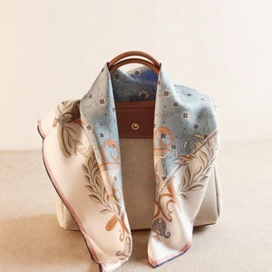 Könnte beinhalten: Ein hellblaues und cremefarbenes Seidentuch mit floralem und Schriftrollenmuster, drapiert über einer beigefarbenen Handtasche mit braunem Ledergriff. Der Schal hat einen rosa und marineblauen Rand.