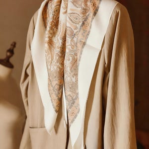 Könnte beinhalten: Ein beiger Blazer mit einem weißen und mit Paisley-Muster versehenen Schal, der darüber drapiert ist. Der Schal hat einen cremefarbenen Rand und ein detailliertes Design in Braun- und Grautönen. Der Blazer hängt auf einem Kleiderständer.