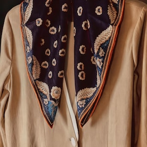 Puede incluir: Una bufanda de seda azul marino con un estampado floral y ribete naranja está drapeada sobre una blazer beige. La bufanda presenta diseños florales blancos y azules. La blazer tiene un botón blanco y un bolsillo.