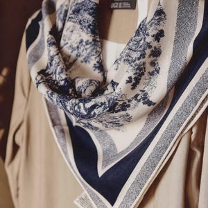 May include: A cream-colored scarf with a blue and white toile pattern of trees and foliage. The scarf has a navy blue border and is draped over a beige garment on a wooden hanger.