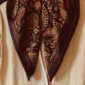 May include: A dark brown scarf featuring a paisley and floral design in red, green, and cream hues. The scarf, with a dark brown border, is draped over a light-coloured blazer.