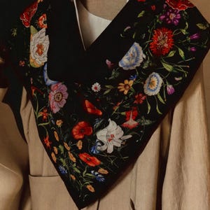 May include: A black scarf with a vibrant floral pattern featuring red poppies, blue cornflowers, and white lilies. The scarf is draped over a beige jacket, with the word "VOGUE" visible on the label.