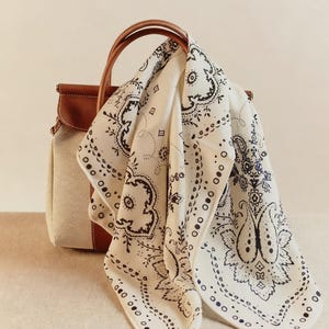May include: A cream-colored handbag with a brown leather handle and trim. A white bandana with a black paisley pattern is draped over the handle. The bandana has a detailed floral design and a dotted border.