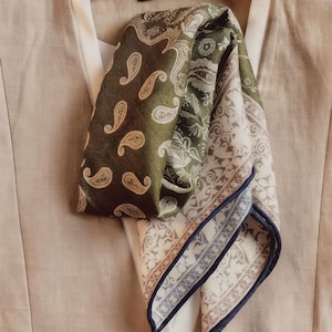 May include: A folded silk scarf featuring paisley and floral designs in olive green, cream, and brown hues. The scarf is draped over a beige garment, with the "VOGUE" label visible. The scarf has a navy blue border.
