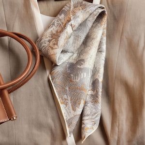 May include: A beige blazer with a white top and a patterned silk scarf. The scarf features a design with beige, grey, and white botanical and architectural elements. A brown leather handbag is visible on the left.