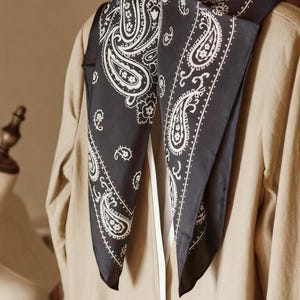 May include: A black paisley bandana with white floral and teardrop designs. The bandana is draped over a tan blazer, showcasing the intricate pattern and contrasting colors. The square scarf is folded into a triangle.