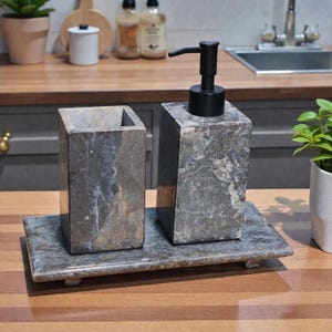 Puede incluir: Un juego de baño de mármol gris que incluye un dispensador de jabón rectangular con una bomba negra, un porta cepillos de dientes y una bandeja rectangular. El juego está hecho de mármol gris con vetas gris oscuro.