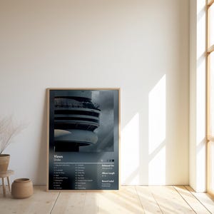 May include: A framed poster featuring a dark image of a building with a circular design. The poster includes the text "Views" and a tracklist. The poster is displayed in a room with a wooden floor, a window, and a small plant.