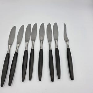May include: A set of eight butter knives with stainless steel blades and dark wooden handles. The knives have a sleek, modern design, perfect for spreading butter or other soft spreads. The handles are approximately 5 inches long.
