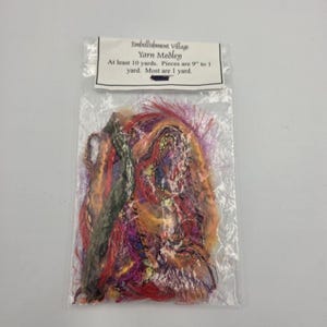May include: A clear plastic bag contains a Yarn Medley from Embellishment Village. The yarn is a mix of colors including red, orange, purple, and green. The text on the label says "At least 10 yards. Pieces are 9" to 1 yard. Most are 1 yard."
