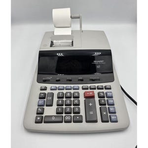 May include: A gray Sharp Compet VX-2650H calculator with a paper roll and a black display. The calculator has a full keypad with number and function keys. The calculator is plugged into a power outlet.