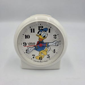 May include: White Lorus Quartz alarm clock featuring a cartoon image of Donald Duck. The clock has a round face with black numerals and hands, and a red second hand. The clock is a vintage Disney collectible.