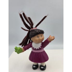 May include: A small plastic doll with dark brown yarn hair styled in a high ponytail, wearing a dark purple dress with a white scalloped collar featuring the letters GPK. The doll has a light pink waistband, black shoes, and holds a green object.