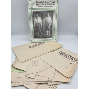 May include: A sewing pattern envelope titled Broadfall Drop Front BREECHES PATTERN with waist sizes 30 thru 42 included, featuring a black and white photograph of two individuals wearing breeches, accompanied by several tan paper pattern pieces.