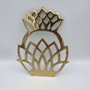 Könnte beinhalten: Ein goldfarbener Metall-Topfuntersetzer mit Distel-Design. Der Untersetzer hat ein detailliertes filigranes Muster aus Rauten und Blütenblättern, das ein dekoratives und funktionelles Küchenzubehör schafft. Das Design ist symmetrisch.