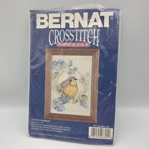 May include: A cross-stitch kit featuring a framed design of a Canada Warbler bird surrounded by blue flowers. The kit includes 14-count Aida fabric, chart, needle, directions, and floss. The package reads "BERNAT CROSSTITCH". The finished design is approximately 5" x 7".