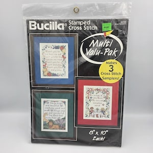 May include: A Bucilla Stamped Cross Stitch Multi Valu-Pak containing materials to make 3 cross stitch samplers. The packaging displays three finished samplers in blue, red, and green frames, each measuring 20.32 cm by 25.4 cm.