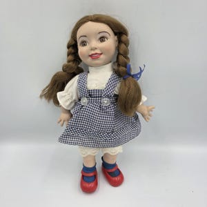 May include: A vintage doll with brown hair styled in two braids, wearing a white blouse, a blue and white checkered dress, and red shoes. The doll has a smiling expression and is a collectible item.