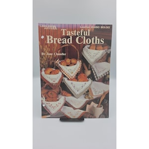 May include: A craft instruction booklet titled Tasteful Bread Cloths by Jane Chandler, Leisure Arts Leaflet 2450. The cover features photographs of various white fabric bread cloths with embroidered designs, displayed in baskets with bread rolls.