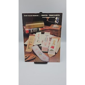 May include: A Mary Ellen Designs Novel Thoughts Charted Hook Marks Vol. II booklet featuring a collection of cross-stitch patterns for bookmarks. The cover displays several finished stitched bookmarks with various text and illustrations on a wooden surface.