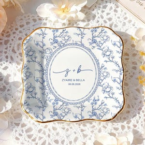 Puede incluir: Un plato cuadrado con diseño floral azul y blanco, borde dorado. El plato tiene los nombres ZYAIRE & BELLA y la fecha 06.08.2026 dentro de un óvalo. El plato está rodeado de flores y sobre un mantel de encaje.