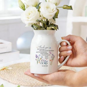 May include: A white ceramic vase with a handle, filled with white roses. The vase has a floral design and text that reads "Thank you for raising the man of my dreams..." The vase is held by a person.