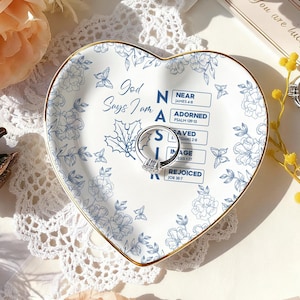 Puede incluir: Un plato de cerámica en forma de corazón con un diseño floral azul y borde dorado. El plato presenta el texto "God Says I Am" y otras palabras. Un anillo de plata con una piedra cuadrada descansa sobre el plato.