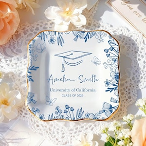 Może przedstawiać: Biała ceramiczna patera na drobiazgi z niebieskim kwiatowym wzorem i złotym obramowaniem. Patera przedstawia ilustrację czapki absolwenta oraz imię "Amelia Smith", a także "University of California" i "CLASS OF 2026".