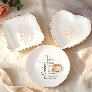 Può includere: Tre portaanelli in ceramica bianca con bordi dorati. Uno è quadrato, uno a forma di cuore e uno rotondo. Ogni piatto ha un disegno a croce e il nome "Sophia" stampato. Il piatto rotondo contiene due anelli d'oro. Una rosa rosa e del verde sullo sfondo.