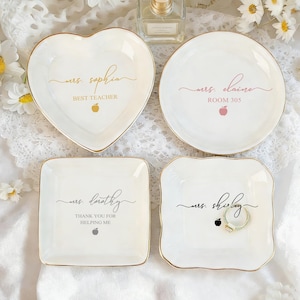 Könnte beinhalten: Vier weiße Keramik-Schmuckschalen mit goldenen Rändern. Eine herzförmige Schale trägt die Aufschrift "Mrs. Sophia, Best Teacher". Weitere Schalen sind rund und quadratisch, mit Namen und Apfelgrafiken. Ein Ring liegt auf einer Schale.