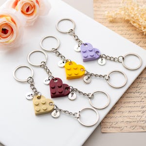 May include: Heart-shaped keychains in various colors, including yellow, purple, and burgundy, made from interlocking blocks. Each keychain has a silver chain and ring, with a small charm bearing a letter. Decorative flowers are in the background.