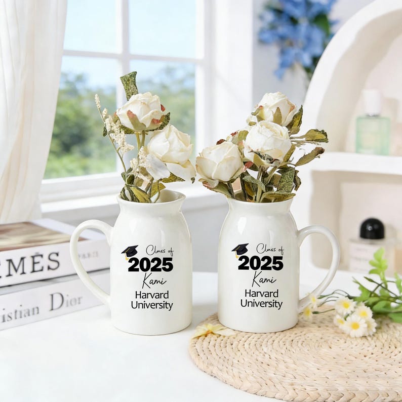 May include: Two white ceramic vases with handles, each holding white roses and greenery. The vases are printed with "Class of 2025 Kami Harvard University" in black. The vases are on a white surface.