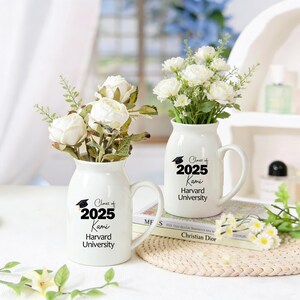 May include: Two white ceramic vases with handles, each holding white flowers. The vases are decorated with the text "Class of 2025 Kami Harvard University" in black. One vase is in front of a book.