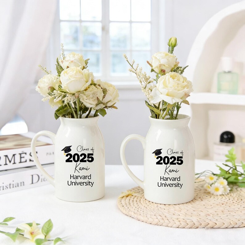 May include: Two white ceramic vases with handles, each holding white flowers. The vases have black text that reads "Class of 2025 Kami Harvard University". The vases are on a white surface.