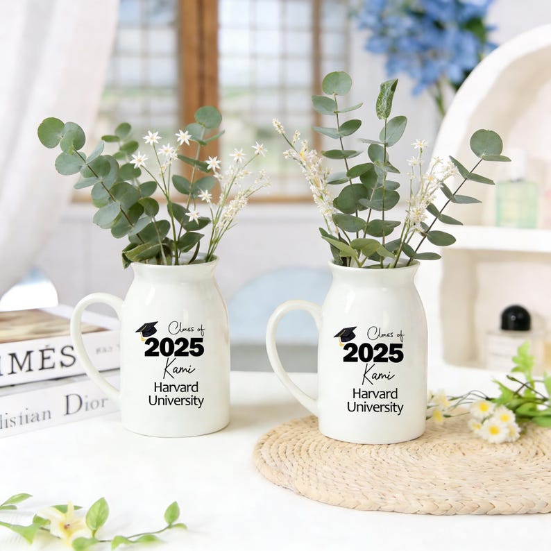 May include: Two white ceramic vases with handles, each displaying the text "Class of 2025 Kami Harvard University" in black. The vases are filled with green eucalyptus and white flowers. A graduation cap graphic is above the text.